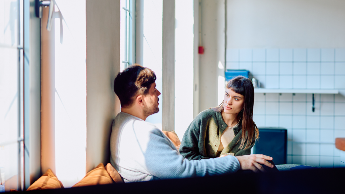 A man and woman sit together in a bright room, having a serious conversation.